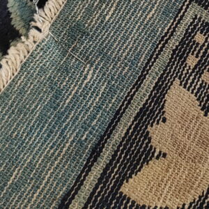 High-pile navy and tan floral rug meeting a teal flatweave backing with a needle stuck in the fringe.
