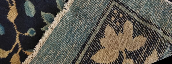 High-pile navy and tan floral rug meeting a teal flatweave backing with a needle stuck in the fringe.