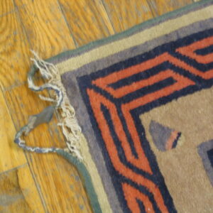 Geometric rug detail showing rust and navy stepped borders and worn fringed edges on wooden flooring.