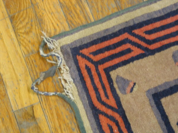 Geometric rug detail showing rust and navy stepped borders and worn fringed edges on wooden flooring.
