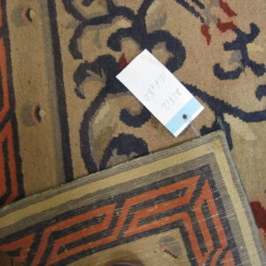 Tan pile rug with dark blue floral motifs and a size tag overlaps an orange geometric flatweave border.