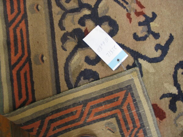Tan pile rug with dark blue floral motifs and a size tag overlaps an orange geometric flatweave border.