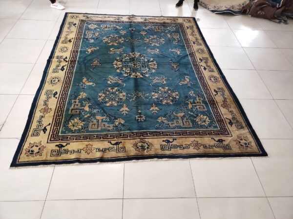 Teal and tan traditional rug with geometric borders and stylized motifs rests on shiny white tiled flooring.