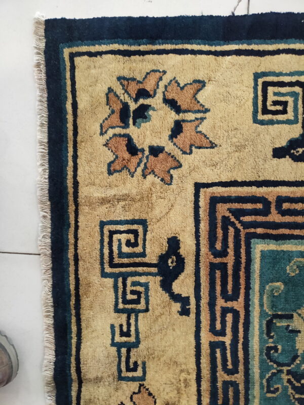 Close-up of a tan rug featuring navy and teal geometric borders and stylized rust-colored floral motifs.