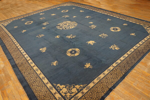 Large square blue rug with tan scattered motifs and a complex geometric border on light wooden flooring.