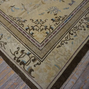 Tan rug with dark brown floral and geometric borders on worn wood flooring.