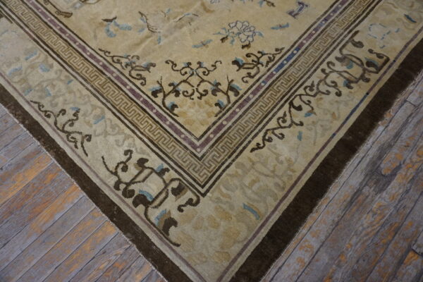 Tan rug with dark brown floral and geometric borders on worn wood flooring.
