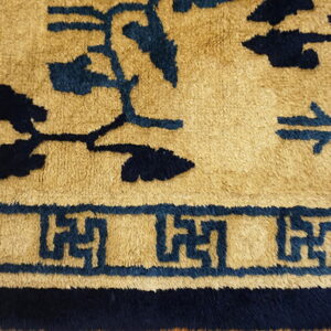 High-pile rug detail showing dark blue scrolling motifs and a fret border on a camel background.