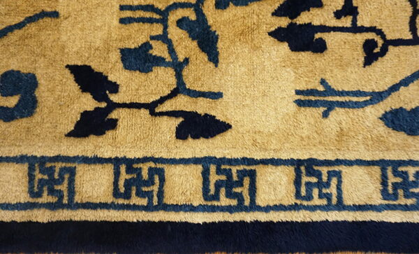 High-pile rug detail showing dark blue scrolling motifs and a fret border on a camel background.