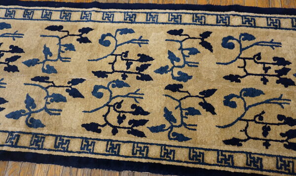 Tan high-pile runner rug featuring dark blue vine patterns and a detailed geometric key border on wood flooring.