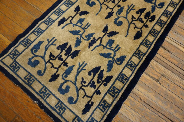 A golden tan rug with dual-tone blue vines and a key pattern border lies on rustic wood flooring.