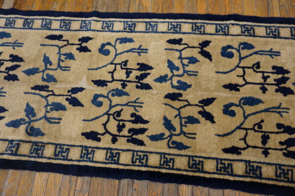 Close-up of a tan rug with dark blue vine patterns and greek key borders on wood planks.