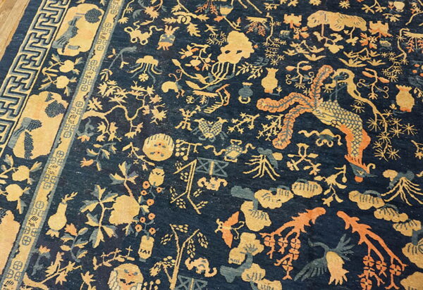 Detailed dark blue rug featuring golden yellow, teal, and orange mythical and floral motifs, with a patterned border on wood floor.