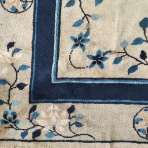 Floral rug corner with a beige field, dark blue border, and blue-pink flowers resting on white tile.