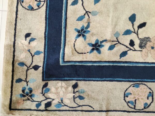 Floral rug corner with a beige field, dark blue border, and blue-pink flowers resting on white tile.
