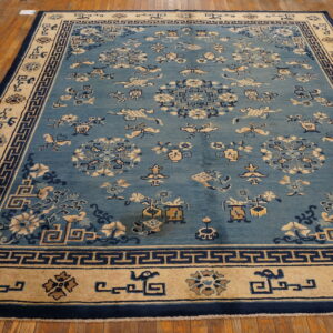 Light blue low-pile rug featuring stylized motifs and navy Greek key borders resting on distressed wooden floors.