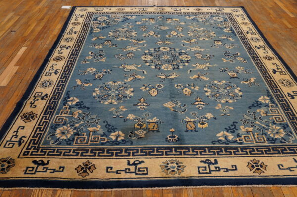 Light blue low-pile rug featuring stylized motifs and navy greek key borders resting on distressed wooden floors.