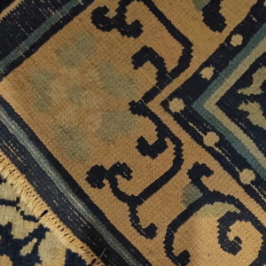 Textured rug detail showing dark blue, tan, and muted green geometric patterns bordered by short fringe.