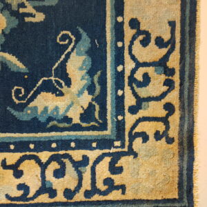 Low-pile rug detail showing a navy field, light blue accents, and tan borders with dark scroll motifs.