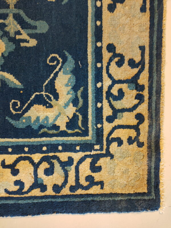 Low-pile rug detail showing a navy field, light blue accents, and tan borders with dark scroll motifs.