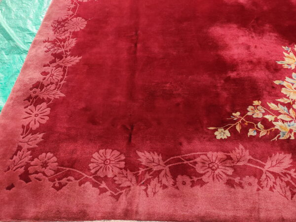 A crimson plush rug features a carved tone-on-tone floral border and a multicolored yellow and blue corner motif.