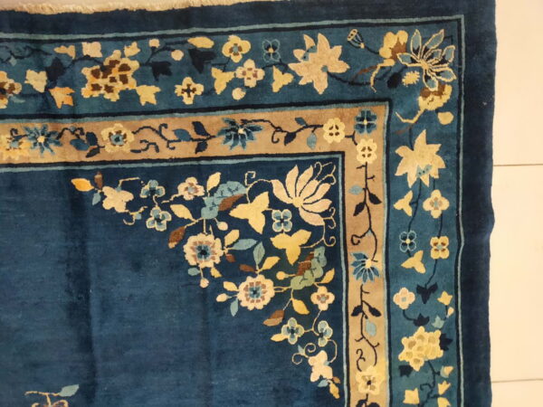 Blue floral motif rug corner resting on a white surface, showing gold, tan, and light blue accents.