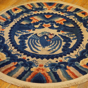 High-pile round rug in blue, beige, and terracotta featuring an ornate stylized bird motif on wood flooring.