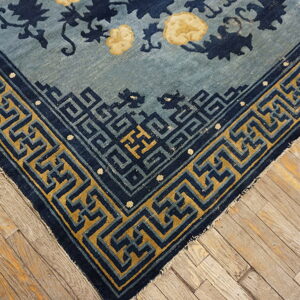 Light blue rug corner with a navy floral pattern and gold geometric key border on wood flooring.
