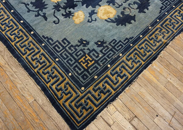 Light blue rug corner with a navy floral pattern and gold geometric key border on wood flooring.