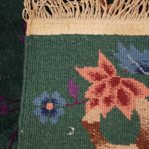 A dark green rug is folded, displaying low pile botanical motifs and cream braided fringe.