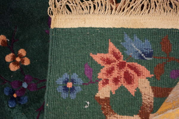 A dark green rug is folded, displaying low pile botanical motifs and cream braided fringe.