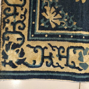 Close-up of a patterned rug corner in deep blue, teal, and tan resting on light tiled floor.
