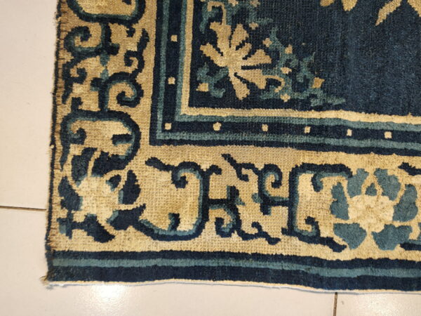 Close-up of a patterned rug corner in deep blue, teal, and tan resting on light tiled floor.