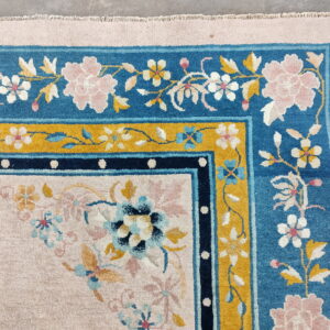 Light pink floral rug with wide borders in bright blue, mustard yellow, and navy blue on concrete floor.