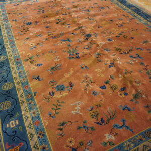 Low pile rug in rust orange with blue florals and a wide patterned border on worn wood flooring.
