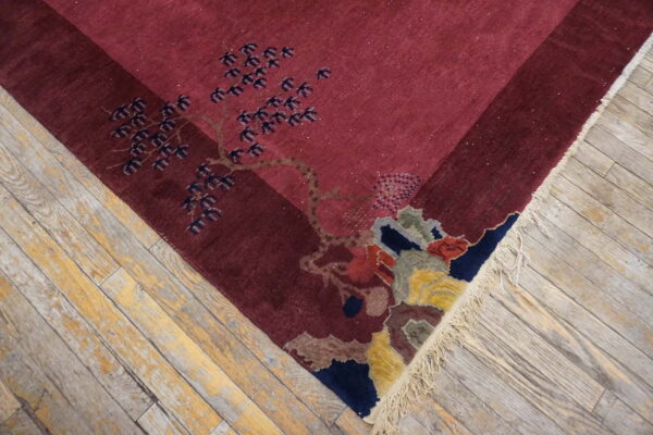 The maroon rug features a multicolored stylized tree pattern and fringe against worn wood flooring.
