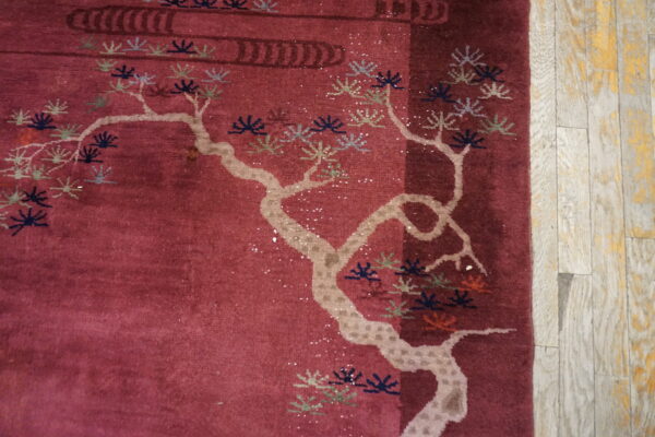 A deep red rug with a light-colored stylized tree design lies on distressed gray and yellow wood planks.