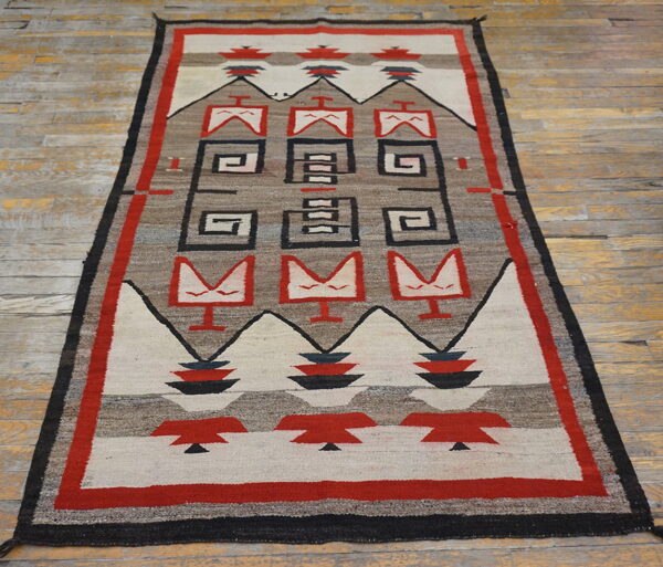 Geometric red, black, and neutral-toned rug with stepped designs and abstract figures on worn wood flooring.