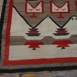 Flat-woven rug with repeating red, cream, black, and brown geometric motifs on worn wood flooring.