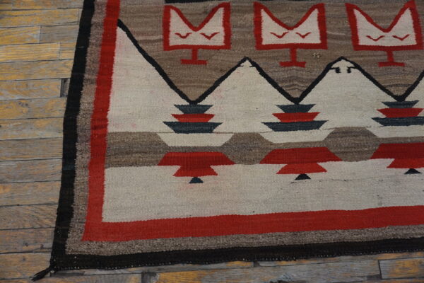 Flat-woven rug with repeating red, cream, black, and brown geometric motifs on worn wood flooring.