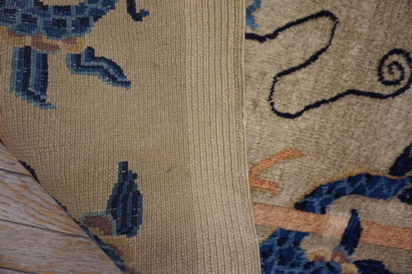 A rug with a plush tan and blue pattern is rolled back, showing its tightly ribbed woven backing on wood flooring.