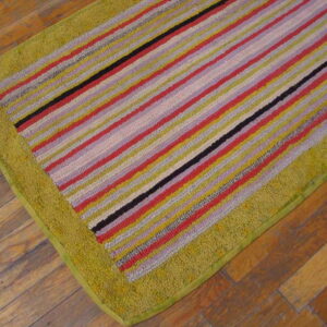 Multicolored striped rug in red, yellow, and black with a high-pile olive green border on wood flooring.
