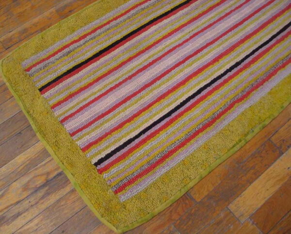 Multicolored striped rug in red, yellow, and black with a high-pile olive green border on wood flooring.