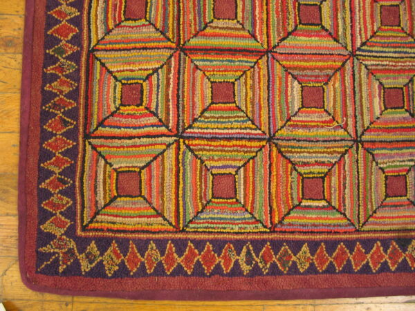 A close-up of a multi-color striped hooked rug featuring geometric blocks and a purple diamond border on wood flooring.