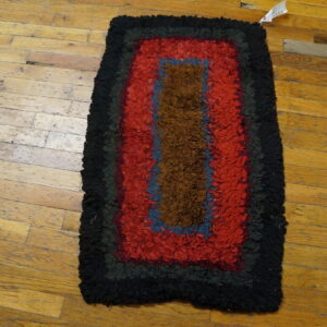 Textured area rug featuring concentric black, red, blue, and brown bands over aged wood flooring.