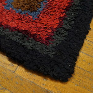 A highly textured, colorful rag rug with geometric blocks of red, black, and blue rests on wood planks.