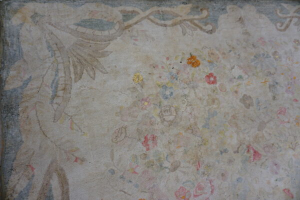Close-up of a low-pile, distressed rug with a faded beige field, formal border, and scattered pale florals.