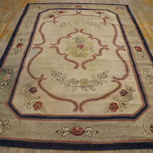 Rectangular beige rug featuring a central floral pattern, scrolling lines, and wide navy and taupe borders on wood planks.