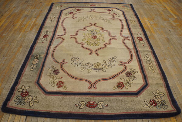 Rectangular beige rug featuring a central floral pattern, scrolling lines, and wide navy and taupe borders on wood planks.