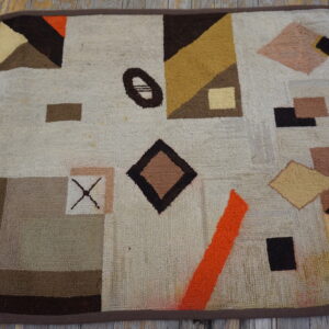 A small, looped rug displays geometric shapes in cream, brown, orange, and gold on wooden flooring.